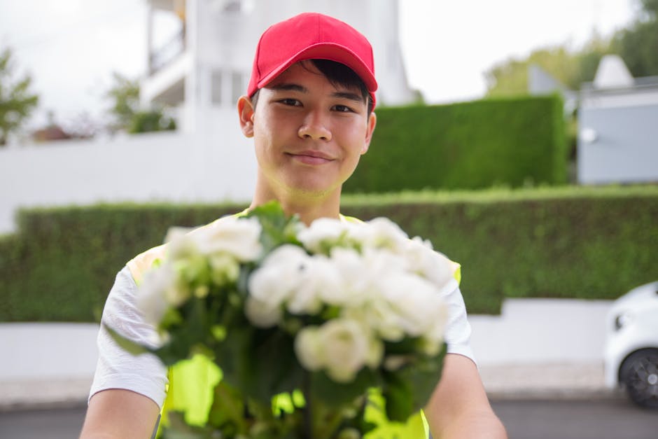 A delivery person holding a bouquet of white flowers with green leaves, dressed in a neon yellow vest and red cap, standing outdoors in front of a modern residential area with neatly trimmed greenery