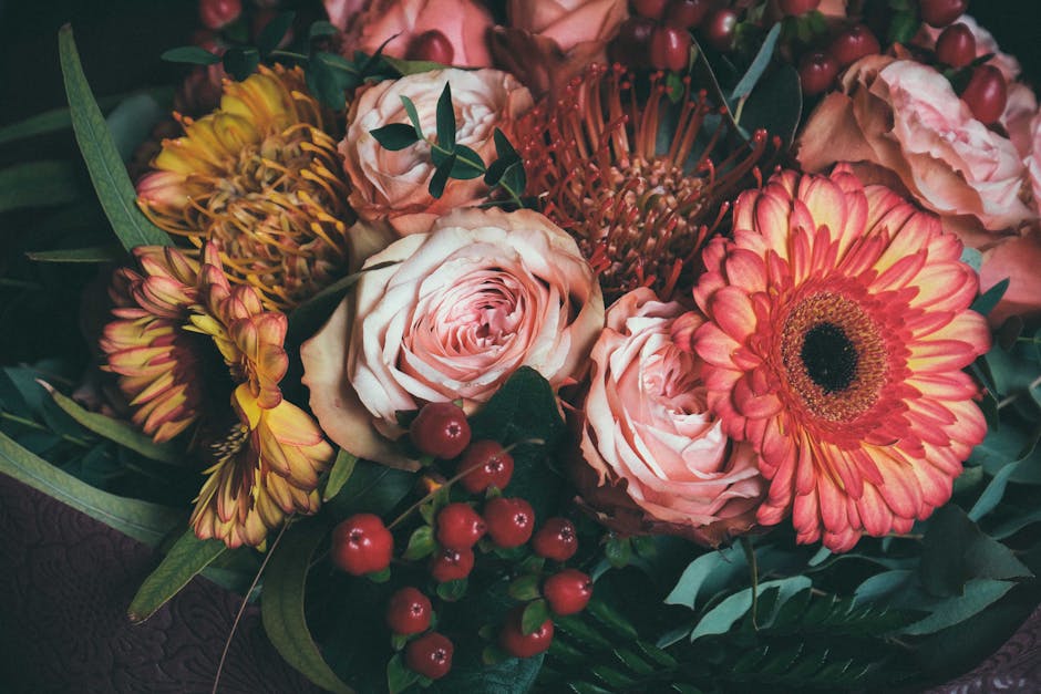 A floral arrangement featuring large, soft-pink roses with delicate, layered petals, accompanied by vibrant pink gerbera daisies with dark centers, and yellow-red chrysanthemums with elongated petals.