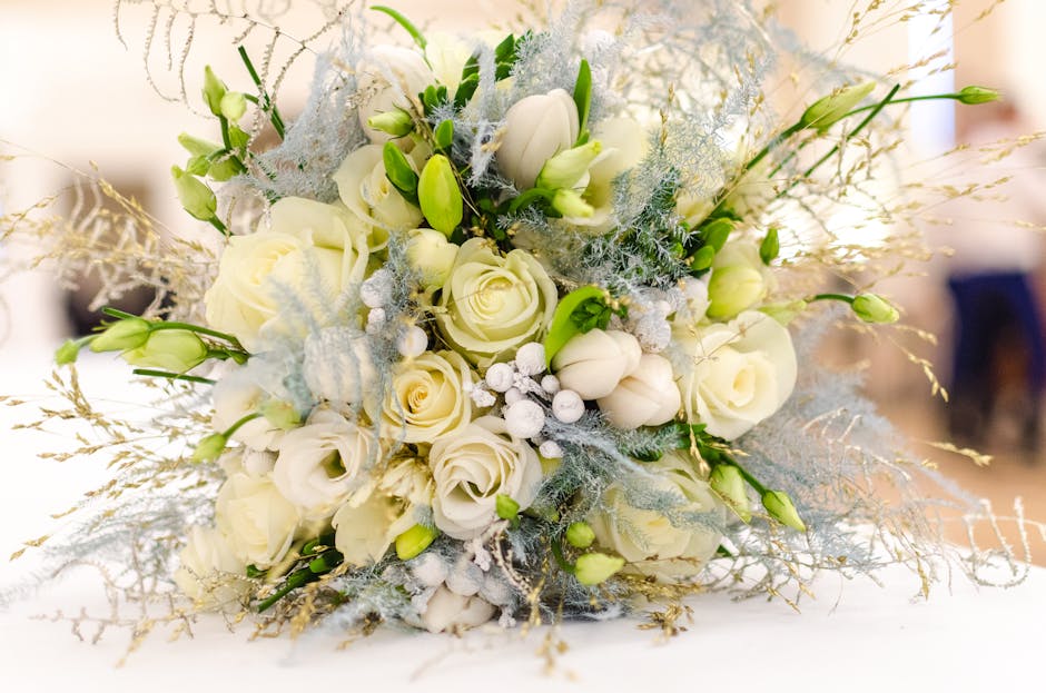 A floral bouquet featuring white roses, eustoma, and delicate white lisianthus arranged with frosted greenery and silver accents, wrapped in elegant pale blue and white tissue paper tied with a satin 