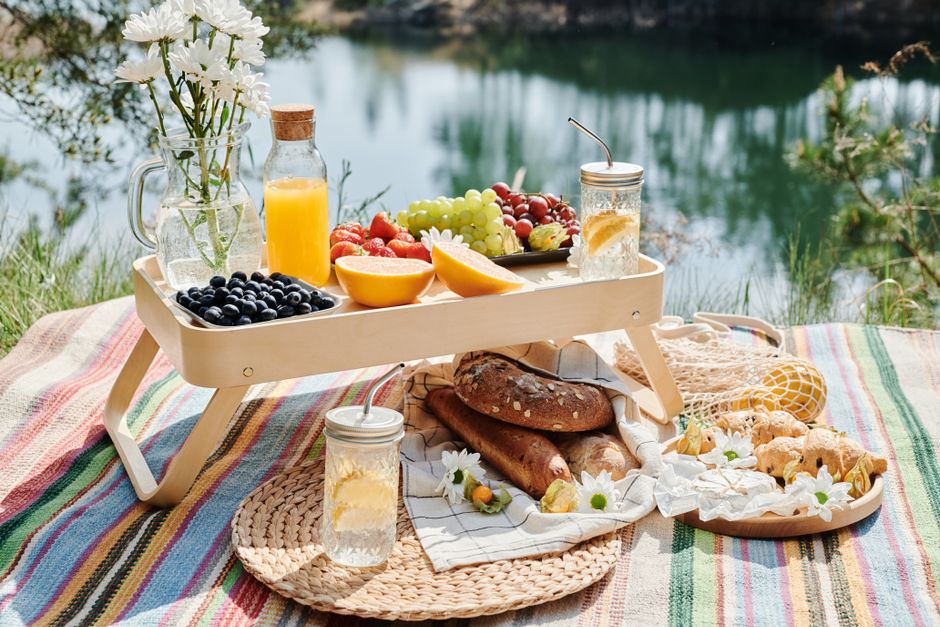 A colorful picnic setup by a tranquil pond features a small light wooden tray with a variety of fresh fruits including green and red grapes, strawberries, blueberries, and halved melons. The arrangeme