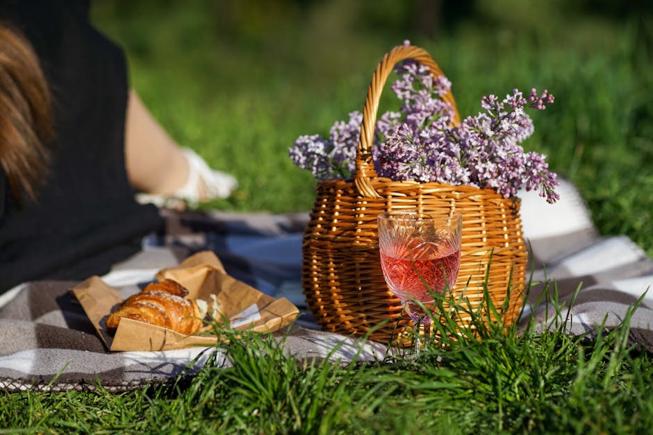 A picnic scene featuring a wicker basket filled with light purple and white heather flowers, placed on a grey and white checkered blanket on lush green grass. Next to the basket, a tall, clear glass w