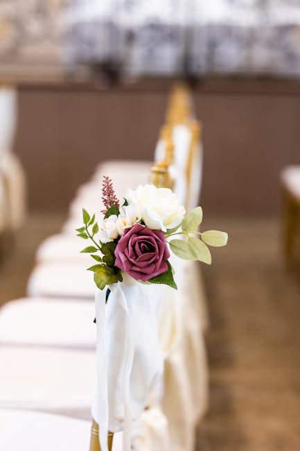 A floral arrangement featuring a bouquet of fresh flowers attached to a white fabric sash draped over a gold-colored banister, with a prominent deep purple rose at the center, complemented by creamy w