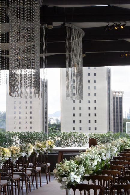 An elegant floral arrangement consisting of white hydrangeas, roses, and greenery decorates a long banquet table set inside a modern venue with a panoramic window overlooking a city skyline with tall