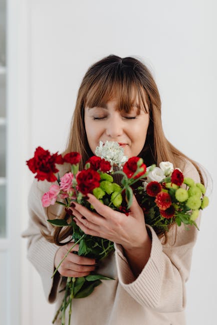 A young woman with brown hair and a gentle smile holds a lush bouquet of fresh flowers, including red carnations, pink roses, white hydrangeas, and green foliage. The bouquet is wrapped in natural bro